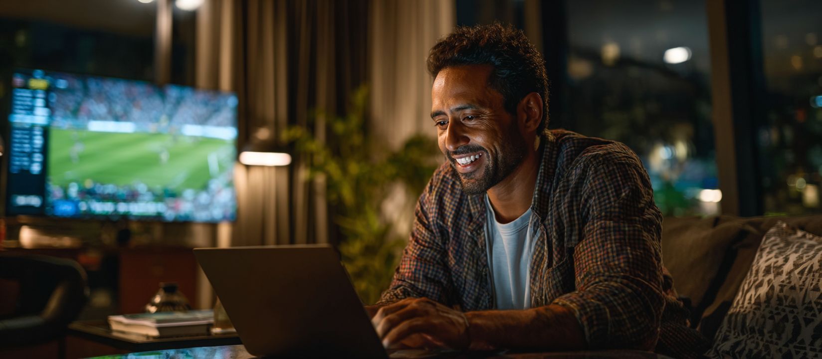 A smiling man is betting on sports online on the 18AA platform while a football match is being broadcast on TV in the background